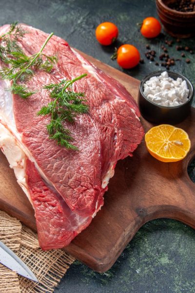 front-view-fresh-raw-red-meats-green-garlics-lemon-salt-brown-wooden-cutting-board-knife-nude-color-towel-tomatoes-pepper-dark-color-background